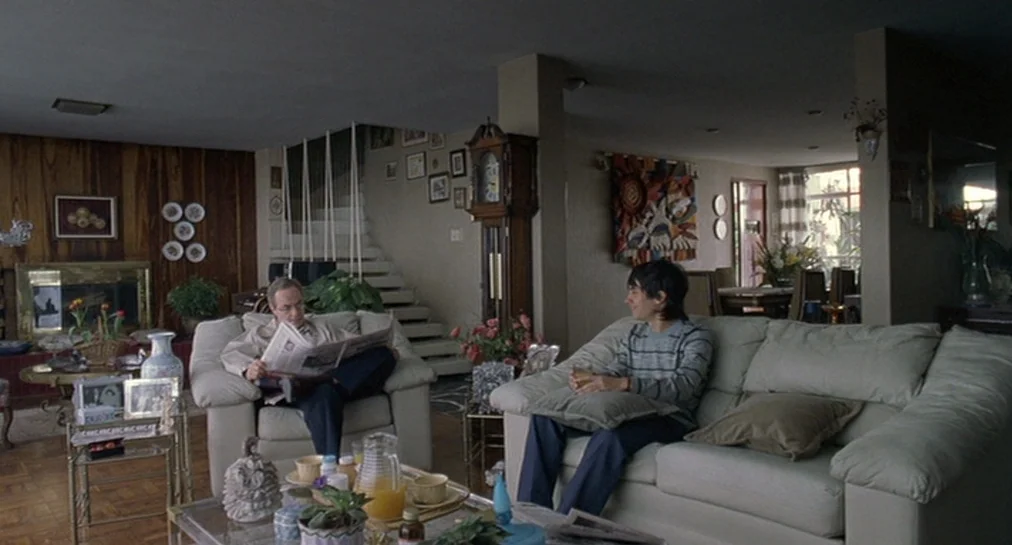 Y Tu Mamá También (2001) movie still: wide — A well-furnished mid-2000s home interior features wood-paneled walls and a grandfather clock. Two men…