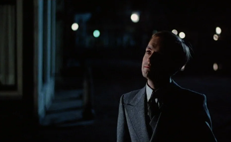 Soldier of Orange (1977) movie still: medium — A dark city street is illuminated by distant blurred street lights at night. A man wearing a suit…