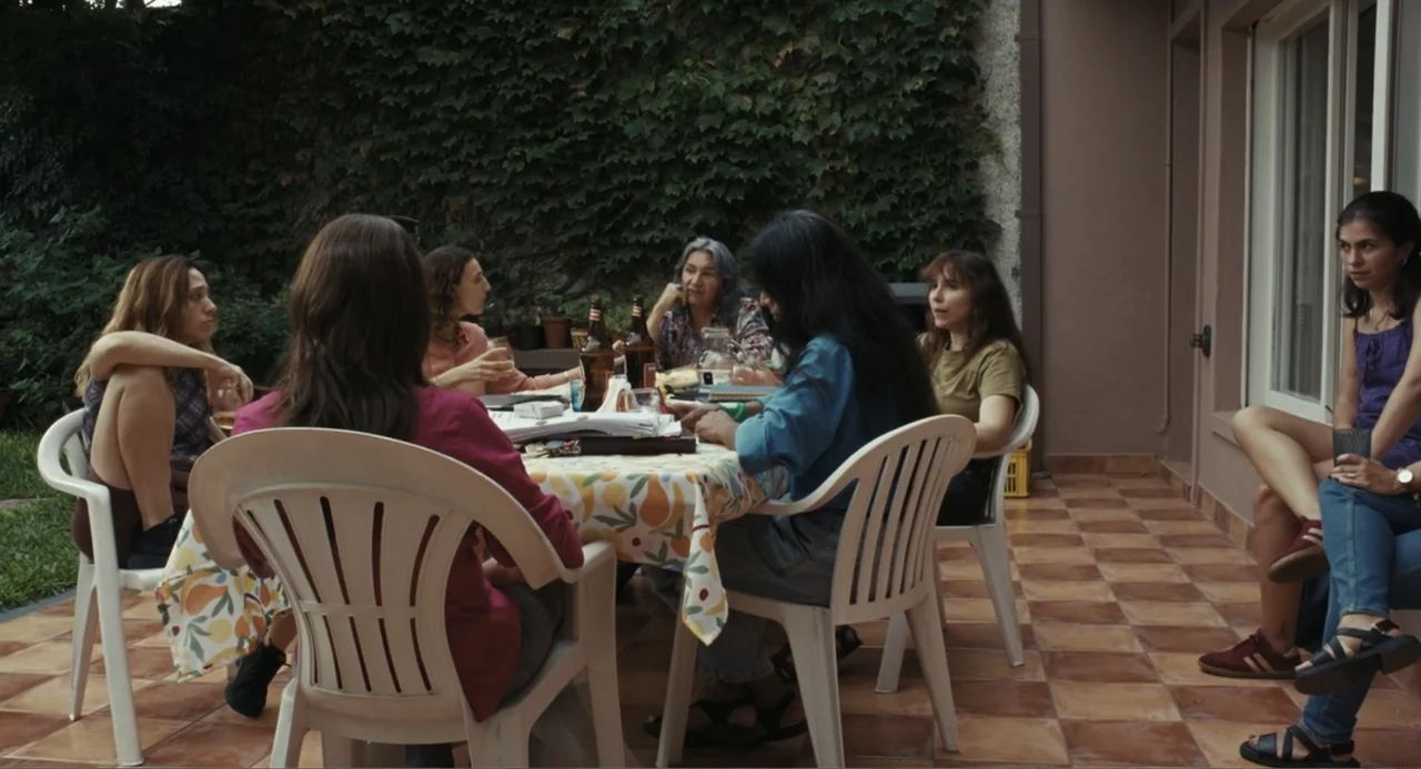 Belén (2025) movie still: fullbody — A terracotta tiled patio features a small table with a floral patterned tablecloth. Six women sit in white…
