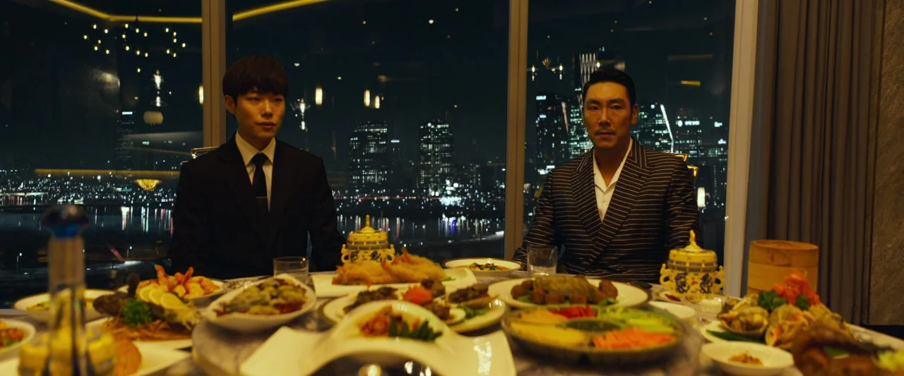 Believer (2018) movie still: medium — A high-rise restaurant at night features a view of city skyscrapers through large windows. Two men sit at a…
