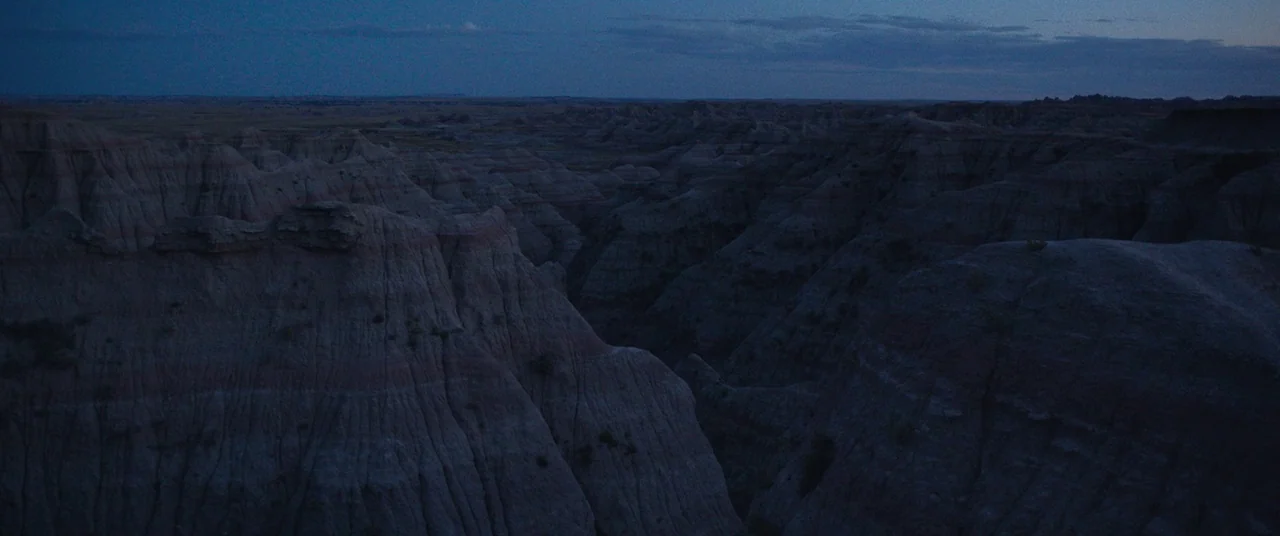The Rider (2017) movie still: establishing — Expansive badlands geological formations feature deep ridges and valleys under a dim twilight sky. No…