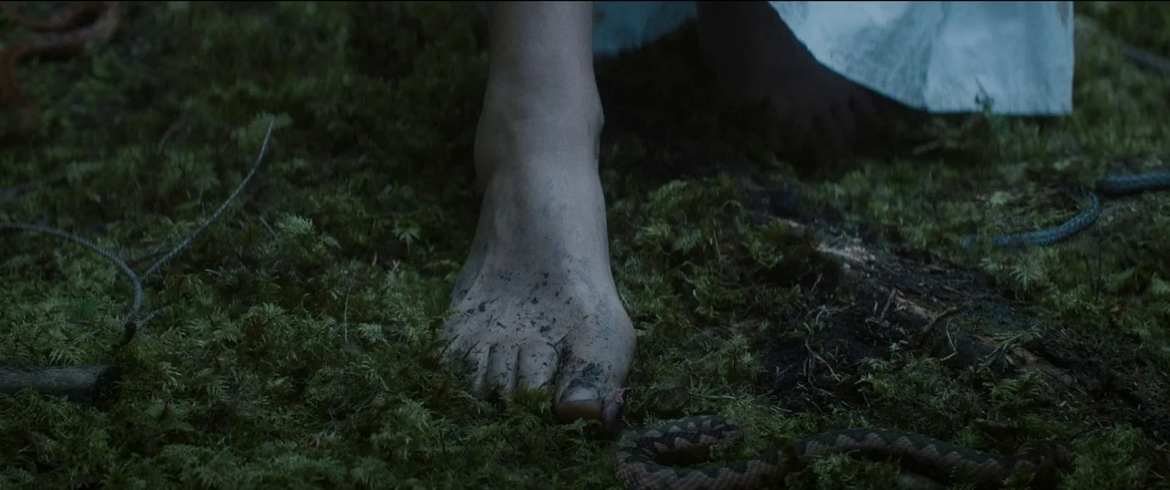 Nightsiren (2022) movie still: closeup — A bare foot covered in dirt stands on a damp mossy forest ground. A small patterned snake lies partially…