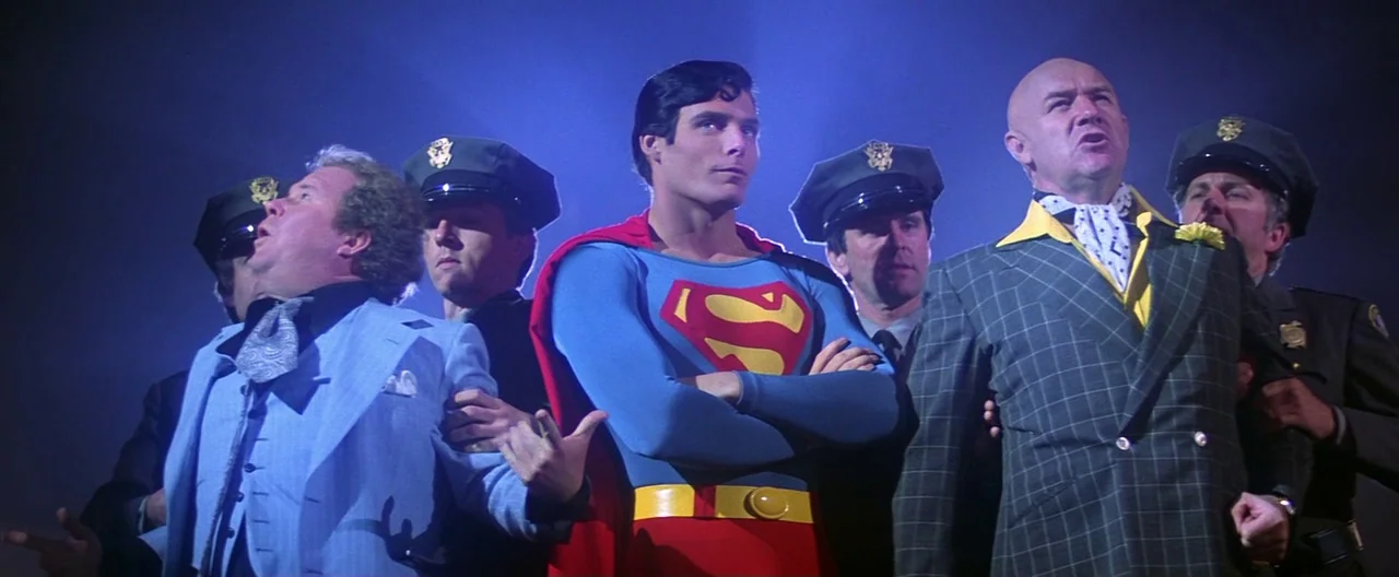 Superman (1978) movie still: medium — A group of men stands together at night under an intense blue sky. Superman holds a center position flanked by…