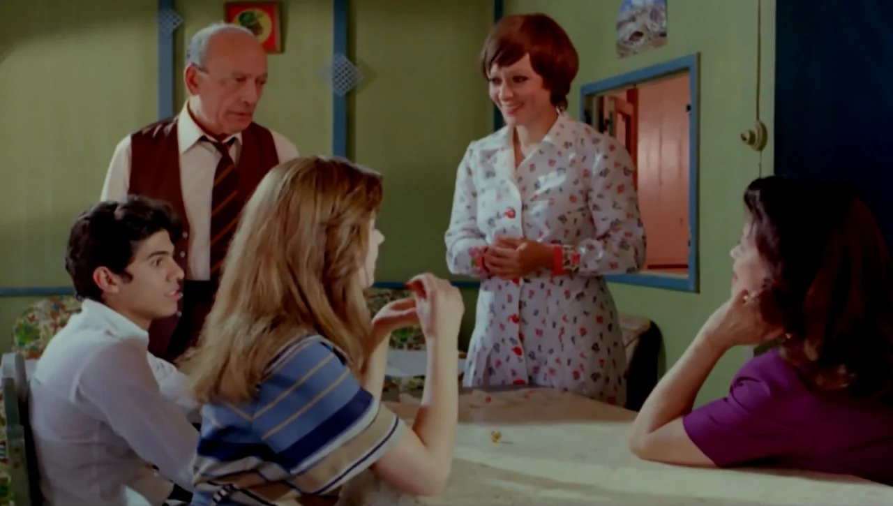 Alexandria…Why? (1979) movie still: medium — A green-walled room with a wooden dining table features small framed wall art. Four individuals are…