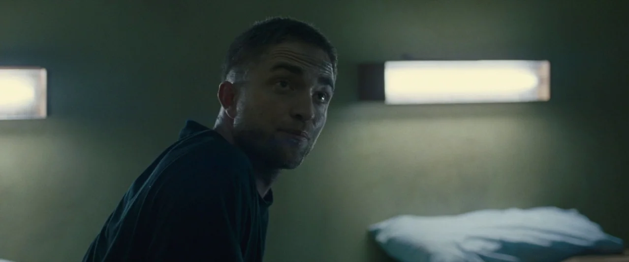 The Rover (2014) movie still: medium — A dimly lit room features concrete walls and two horizontal fluorescent lamps on the wall. A man sits at the…