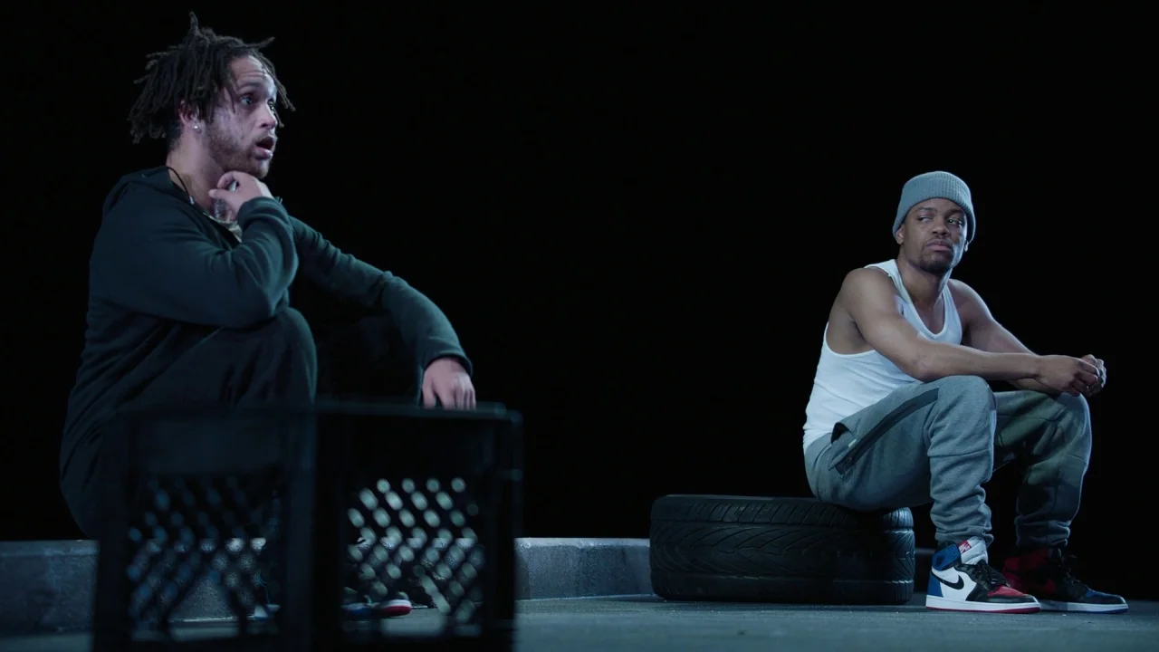 Pass Over (2018) movie still: fullbody — Two men sit on a flat surface in the dark of night. One man perches on a black plastic crate while another…
