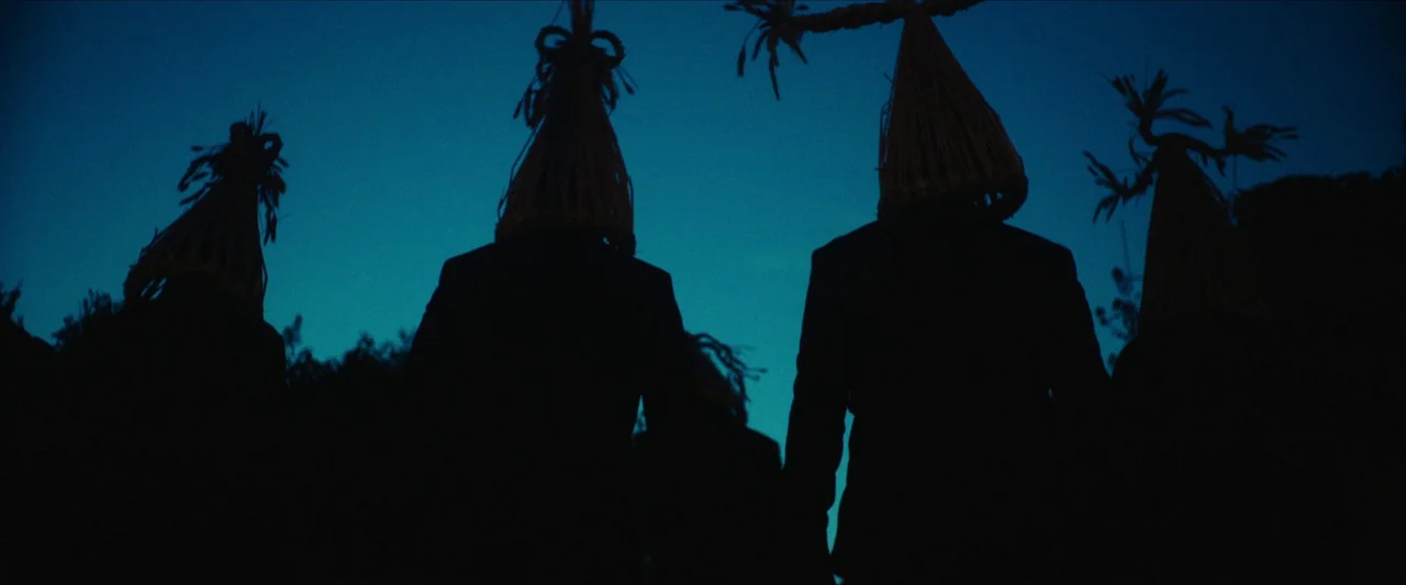Fréwaka (2024) movie still: medium — A group of figures in suits wears pointed straw headdresses in a dark open space. The subjects walk away from…