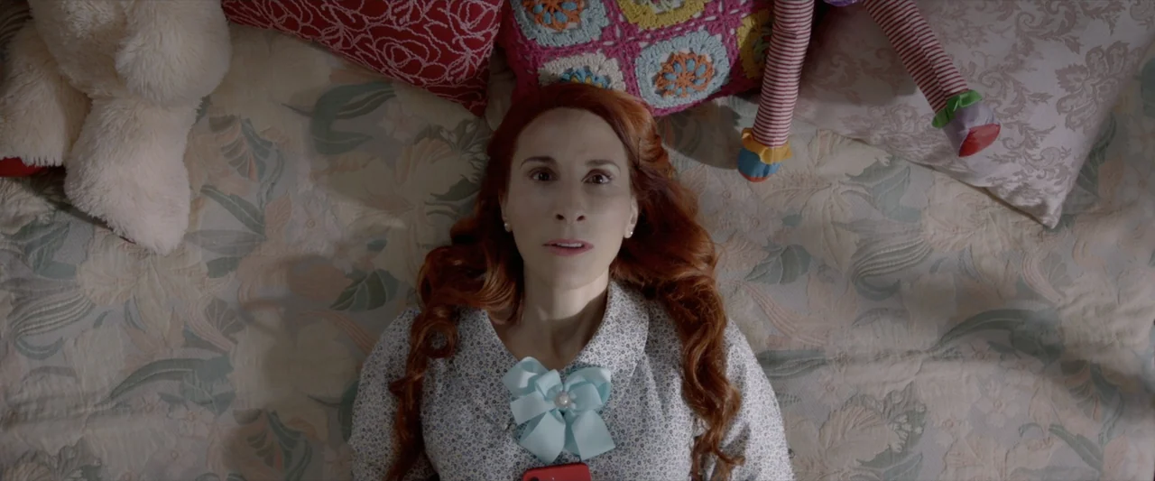 Like or Die (2023) movie still: medium — A woman lies on a floral patterned duvet surrounded by plush toys. She stares directly toward the camera…
