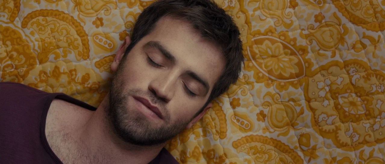 Road Games (2015) movie still: closeup — A man lies on a yellow patterned quilt with a floral paisley print. The subject has his eyes closed and is…