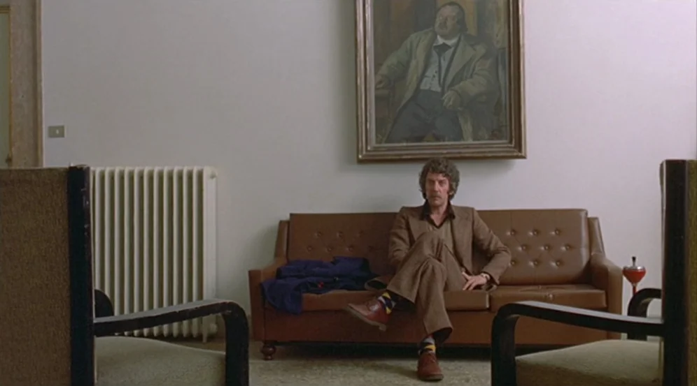 Don’t Look Now (1973) movie still: wide — A minimalist room contains a tan leather sofa, a radiator, and a wall painting. A man in a brown suit sits…