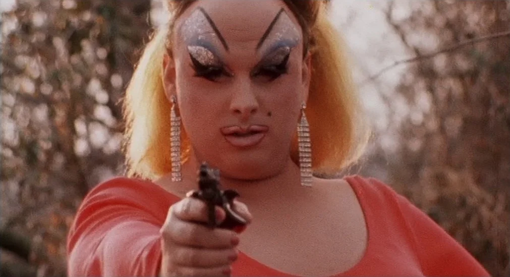 Pink Flamingos (1972) movie still: closeup — A person in drag makeup stands outside near blurred, leafless trees. They hold a small black handgun…