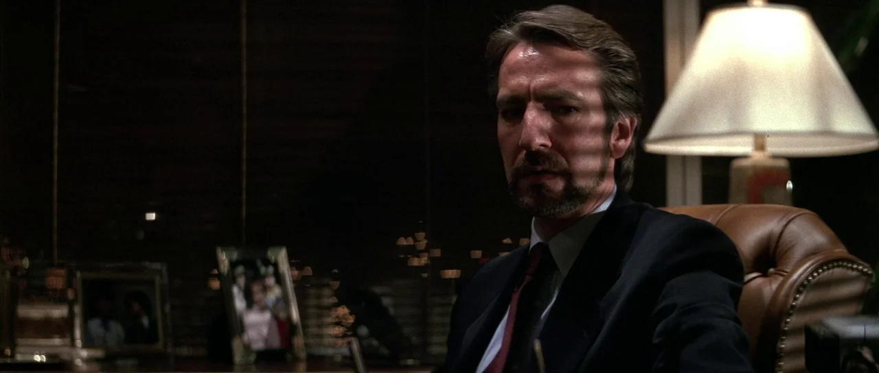 Die Hard (1988) movie still: medium — A dark, wood-paneled office environment with blinds casting shadows over the desk area. A man in a dark suit…