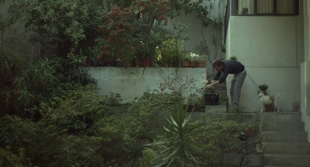 Beginners (2010) movie still: wide — A man stands on concrete steps next to a white wall in a overgrown space. He adjusts a plant pot while holding…