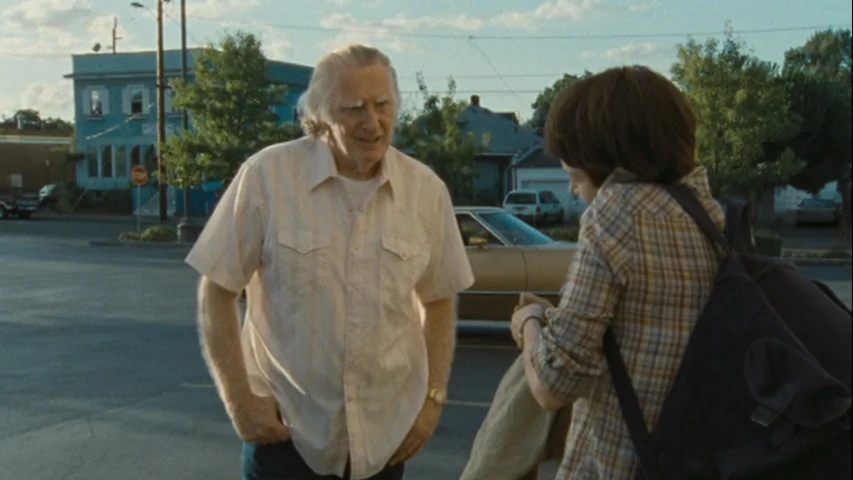 Wendy and Lucy (2008) movie still: medium — A suburban parking lot with a multi-story building and parked cars in the background. An older man…