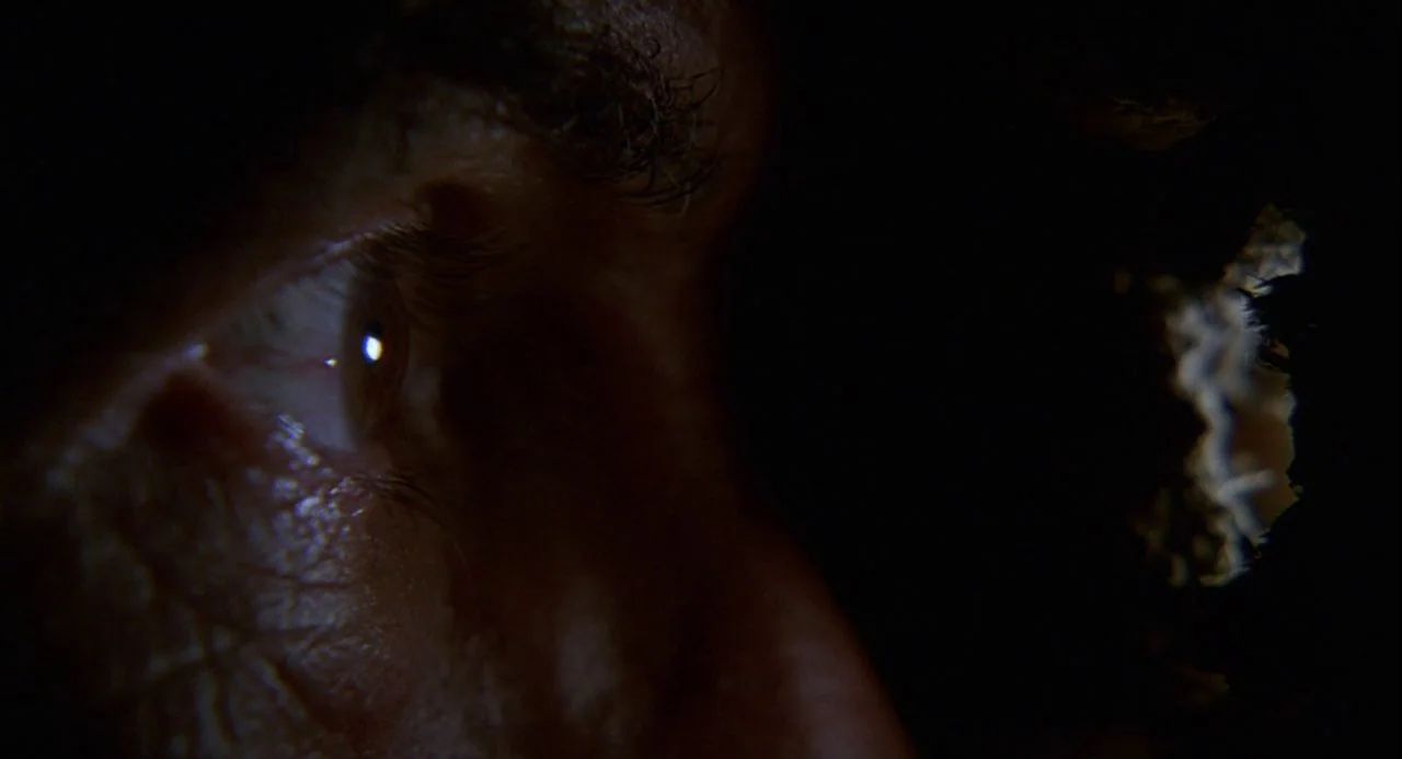 Psycho III (1986) movie still: closeup — A dark, cramped space frames a single human eye pressed against a small opening. The eye looks through a…
