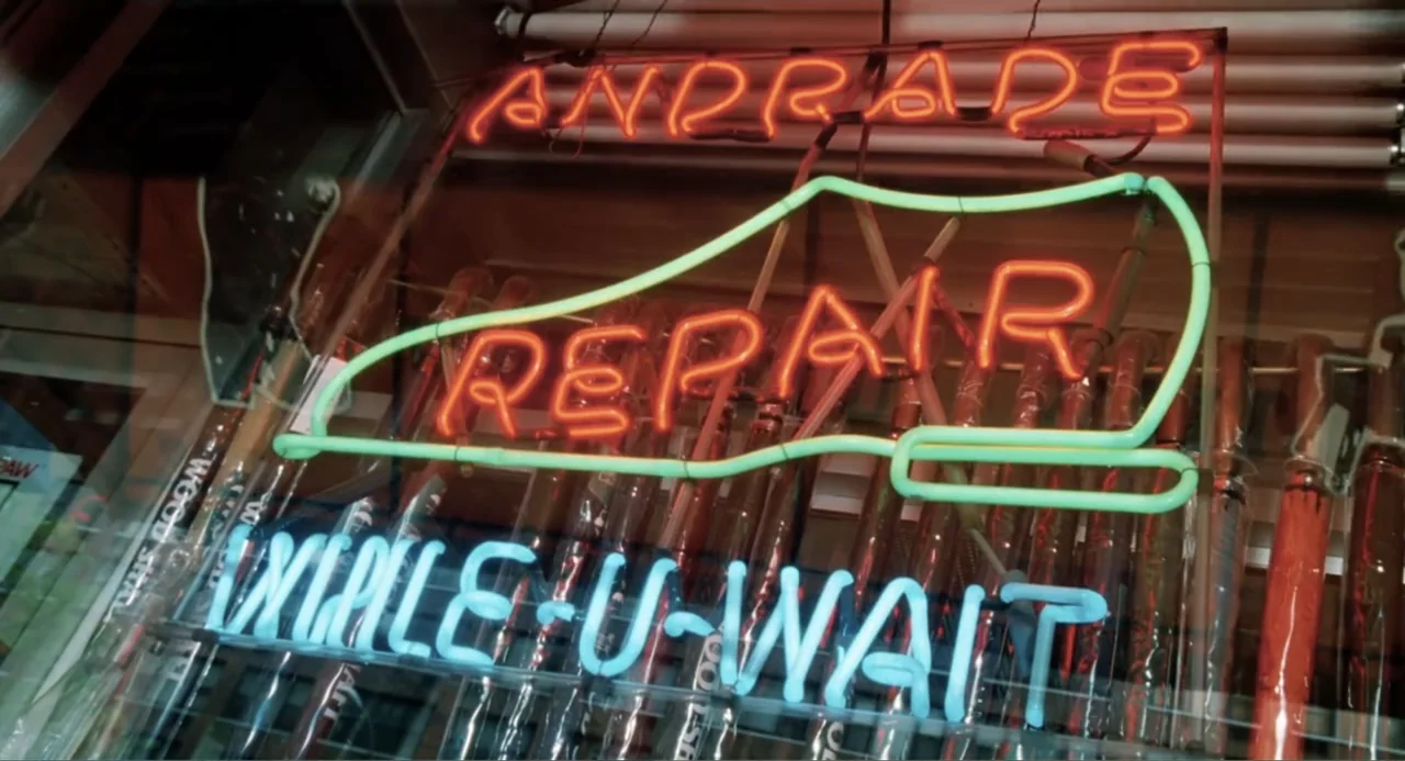 You've Got Mail (1998) movie still: closeup — A neon sign in a storefront window advertises shoe repair services at night. The glass reflects the…