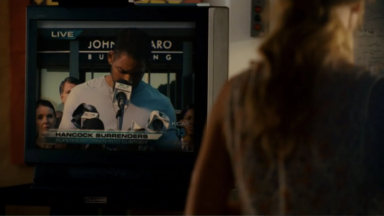 Hancock (2008) movie still: medium — A living room interior with a large television displaying a live news broadcast. A person in the foreground…