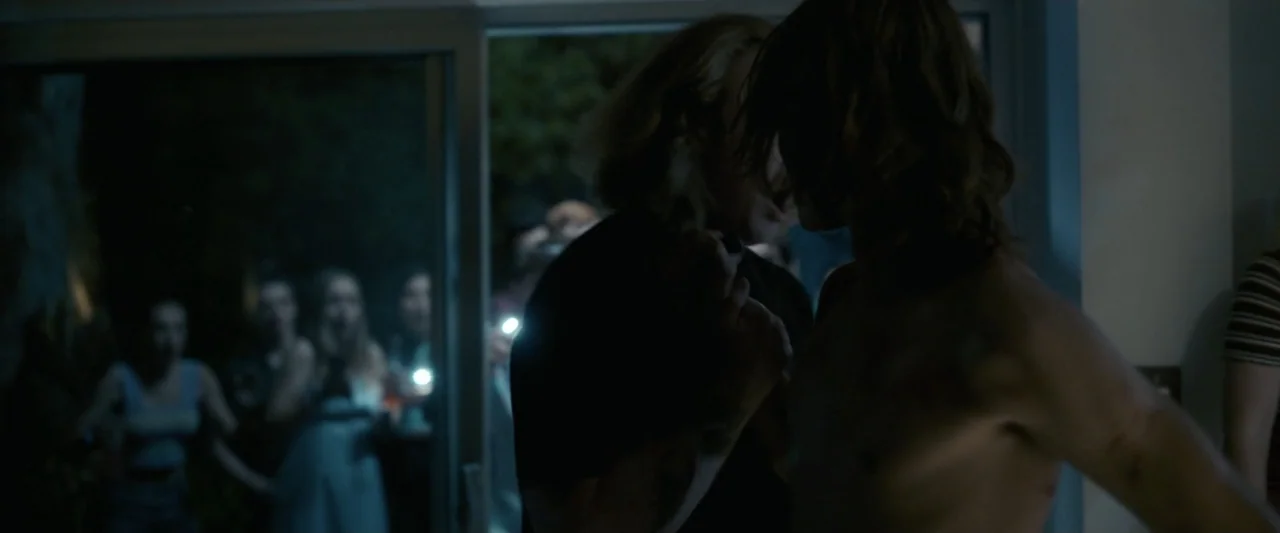 Talk to Me (2023) movie still: medium — A nighttime gathering occurs in a dimly lit space near an open glass door. Two men stand in the foreground…