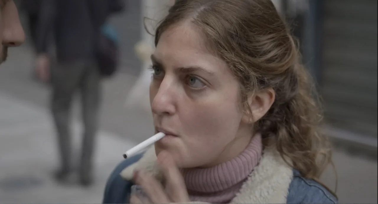 An Odd Turn (2024) movie still: closeup — An outdoor city sidewalk with blurred urban surroundings in the soft background. A young woman holds a lit…
