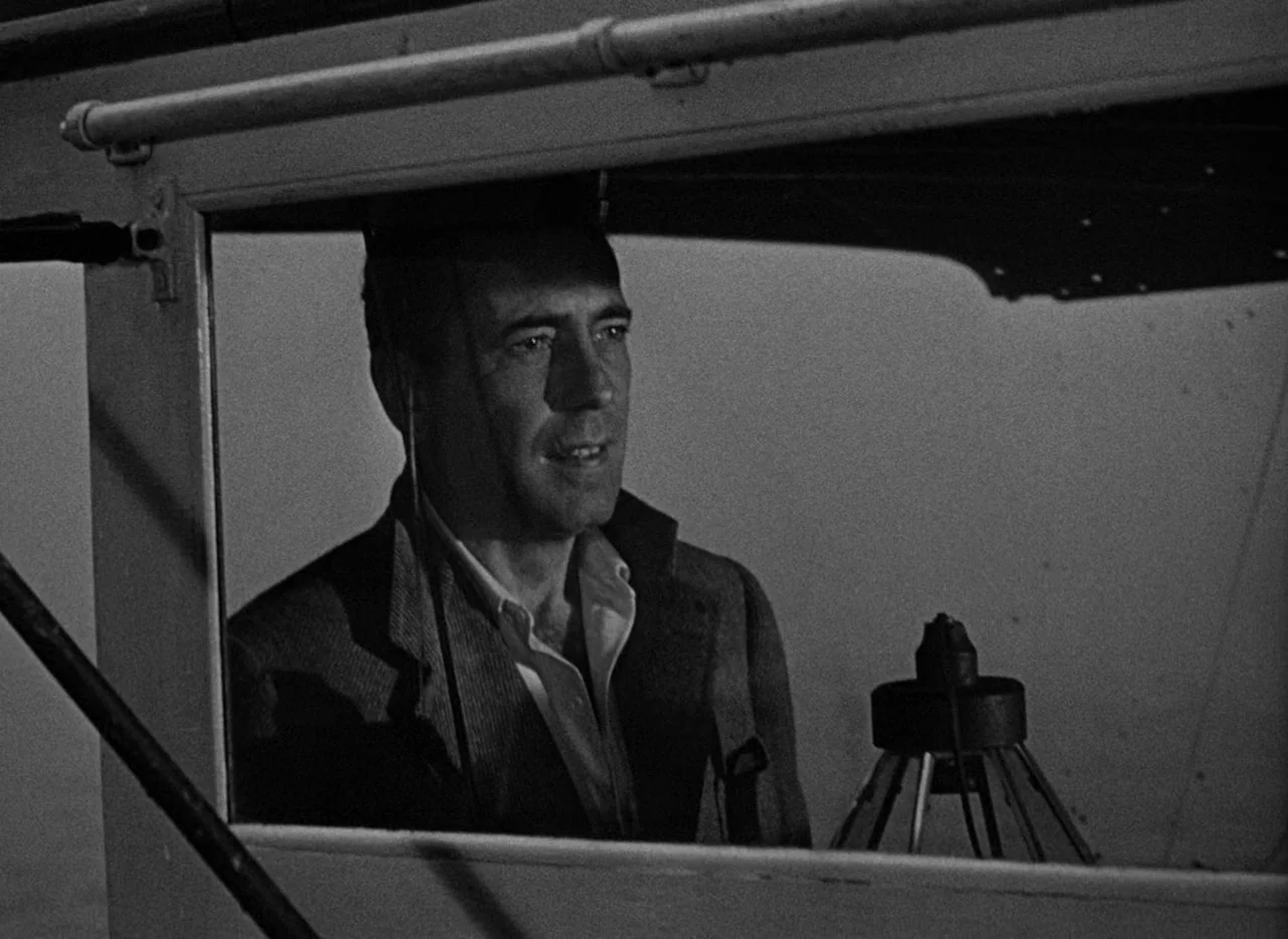 Key Largo (1948) movie still: medium — A dark boat cabin frame obscures the view of the man sitting within. The man looks out through a window while…