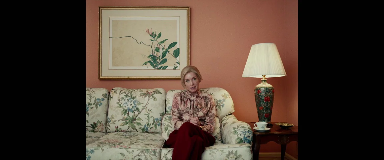 I, Tonya (2017) movie still: medium — A living room features coral-colored walls with a botanical print in a wooden frame. A woman with blonde hair…