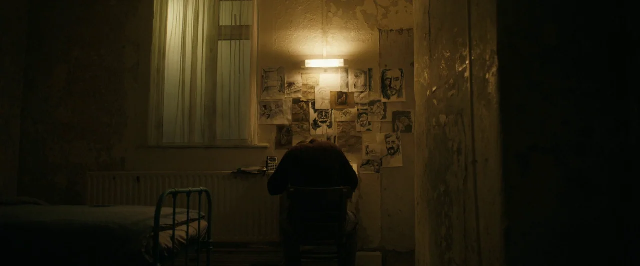 Oddity (2024) movie still: wide — A dimly lit, sparse room contains a bed, a desk, and wallpaper peeling in patches. A lone individual sits at the…