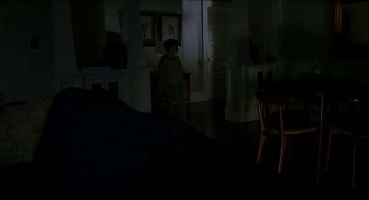 The Ring Two (2005) movie still: medium — A dimly lit room with wooden dining furniture and interior architectural pillars in shadows. A young boy…