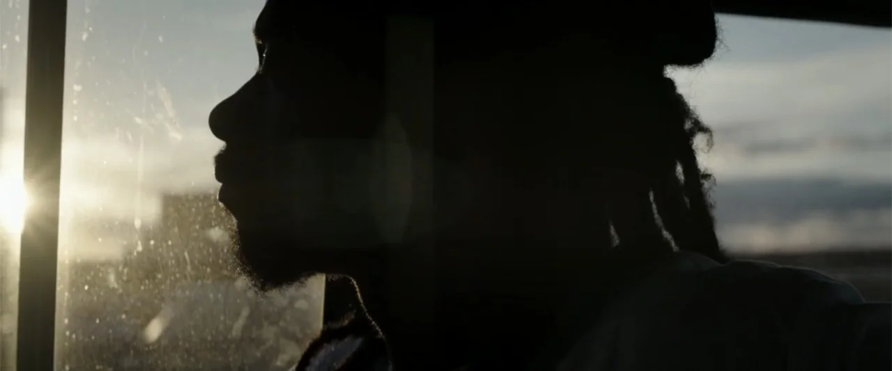Crown Heights (2017) movie still: closeup — A man in dreadlocks sits inside a vehicle looking through a side window. The sun shines brightly against…