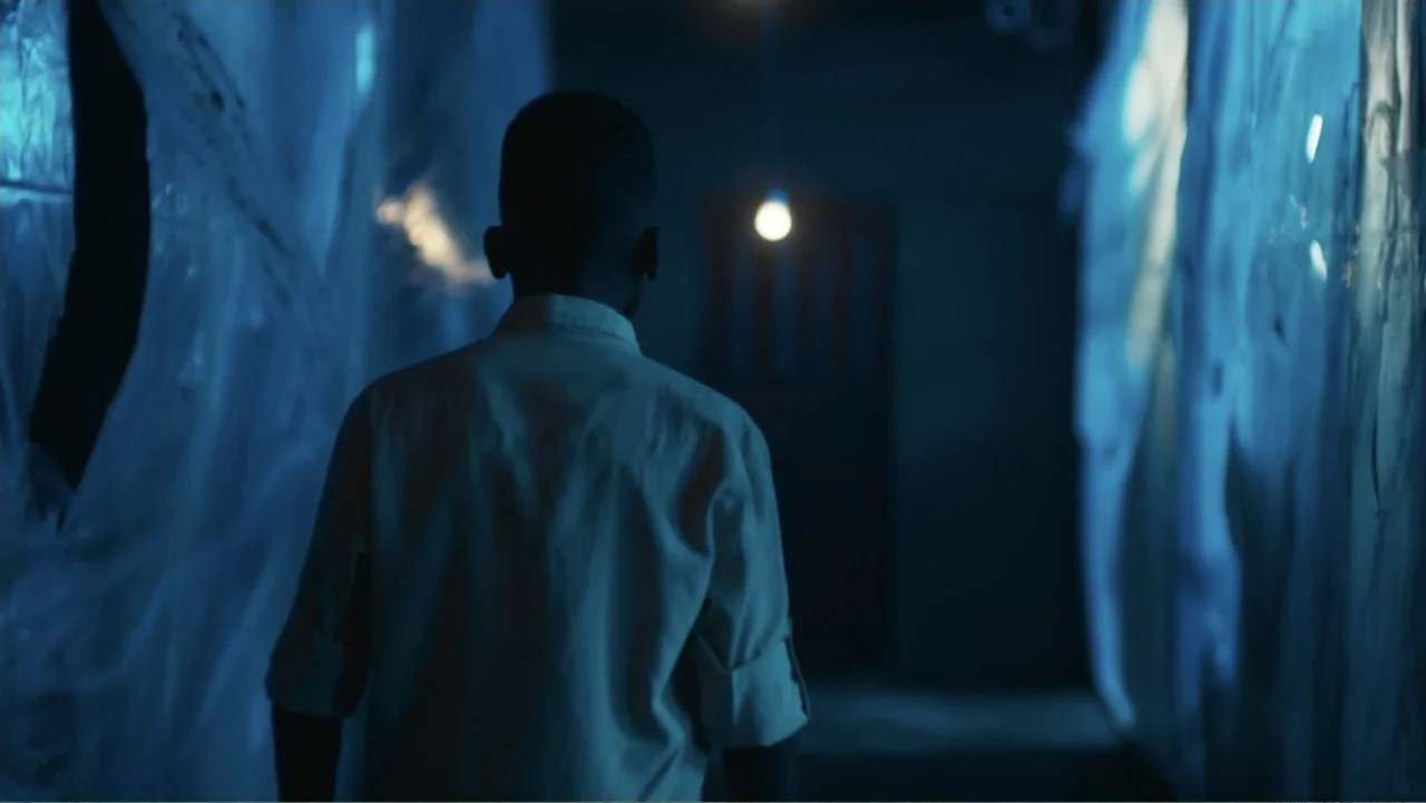 Gangs of Lagos (2023) movie still: medium — A narrow dark corridor is lined with hanging translucent plastic sheets. A man in a light shirt walks…