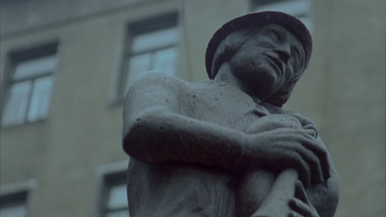 Museum Hours (2012) movie still: medium — A stone sculpture of a man playing a pipe against a grey building facade. The statue is positioned in…