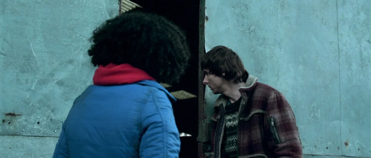 Isolation (2005) movie still: medium — Two individuals stand in front of a weathered, teal-colored corrugated metal wall. They are positioned near a…