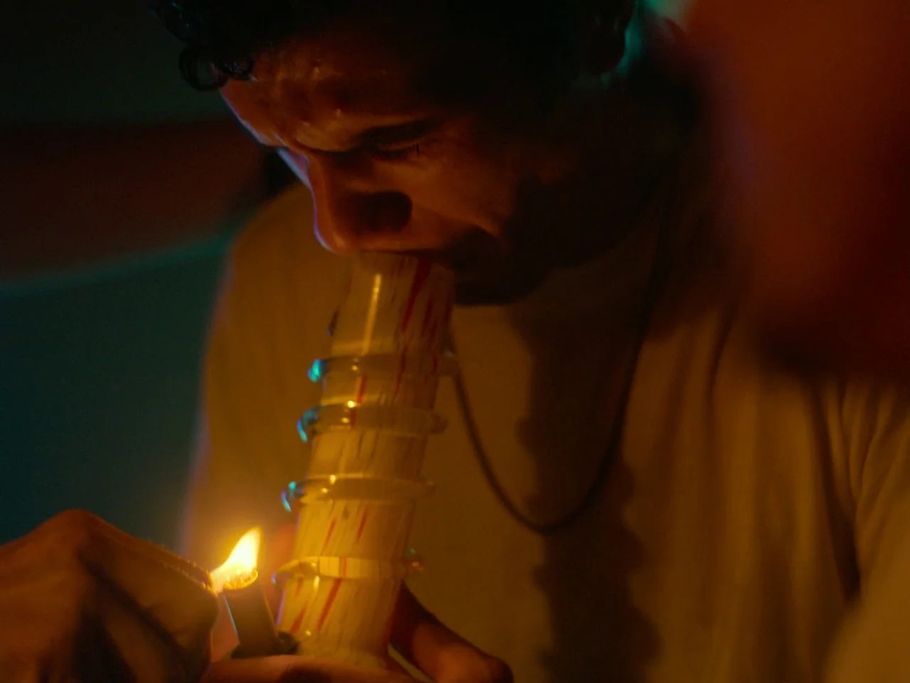 Of An Age (2022) movie still: closeup — A dimly lit room with a person in a beige shirt. The individual is using a lighter to ignite a glass pipe.