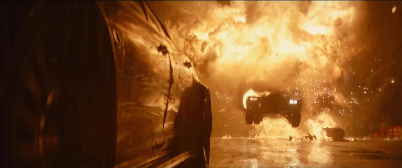 The Batman (2022) movie still: wide — A dark night highway is engulfed by a large, intense fireball explosion. A black sports car leaps through the…