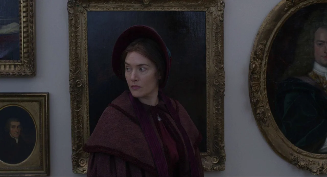 Ammonite (2020) movie still: medium — A woman stands before a wall covered in gilded oil paintings inside a museum. She wears period attire and…