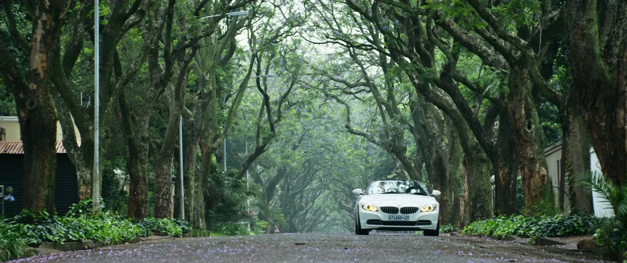 Chappie (2015) movie still: wide — A residential road is lined with tall, mature trees forming a natural archway overhead. A white BMW convertible…