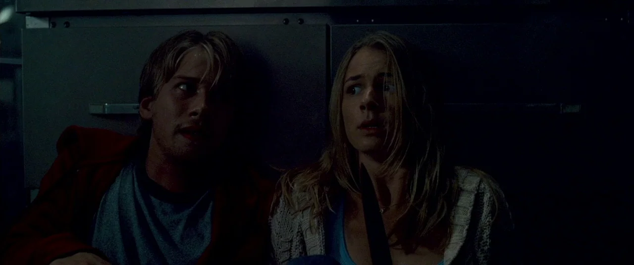 Carriers (2009) movie still: medium — A dark, cramped space with flat metal paneling along the wall behind the subjects. Two young adults sit close…
