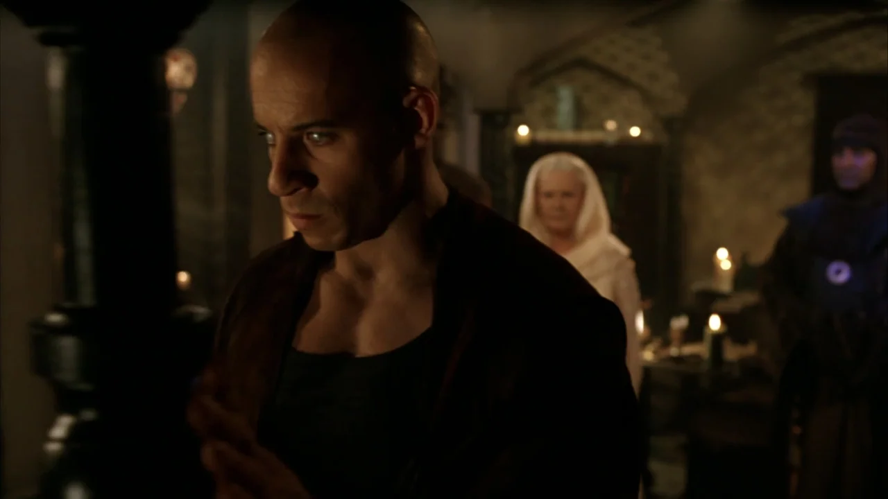 The Chronicles of Riddick (2004) movie still: medium — A stone hall features candlelit backgrounds with blurred figures visible in the distance. A…