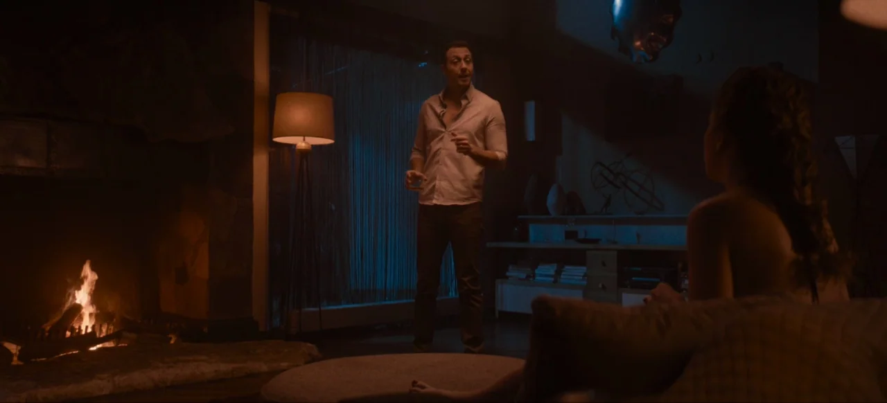 Housewife (2017) movie still: medium — A dimly lit room features a stone fireplace burning with active orange flames. A man stands in the center…