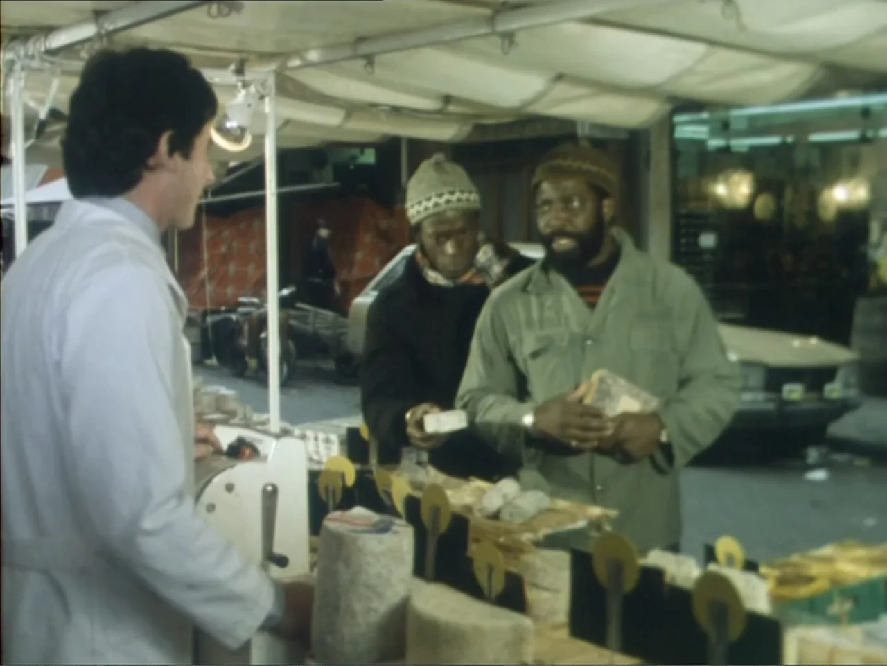 A Dessert for Constance (1981) movie still: medium — A nighttime outdoor market stall displays various goods under a white canvas awning. Two men…