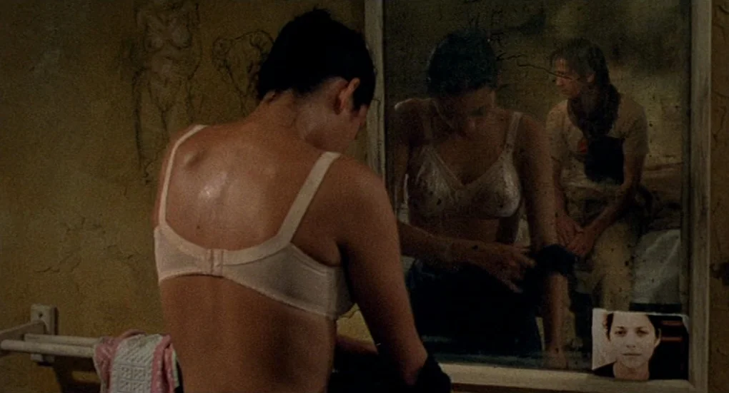 Furia (1999) movie still: medium — A dimly lit room features a worn mirror reflecting a man and woman. The woman stands before the mirror while the…