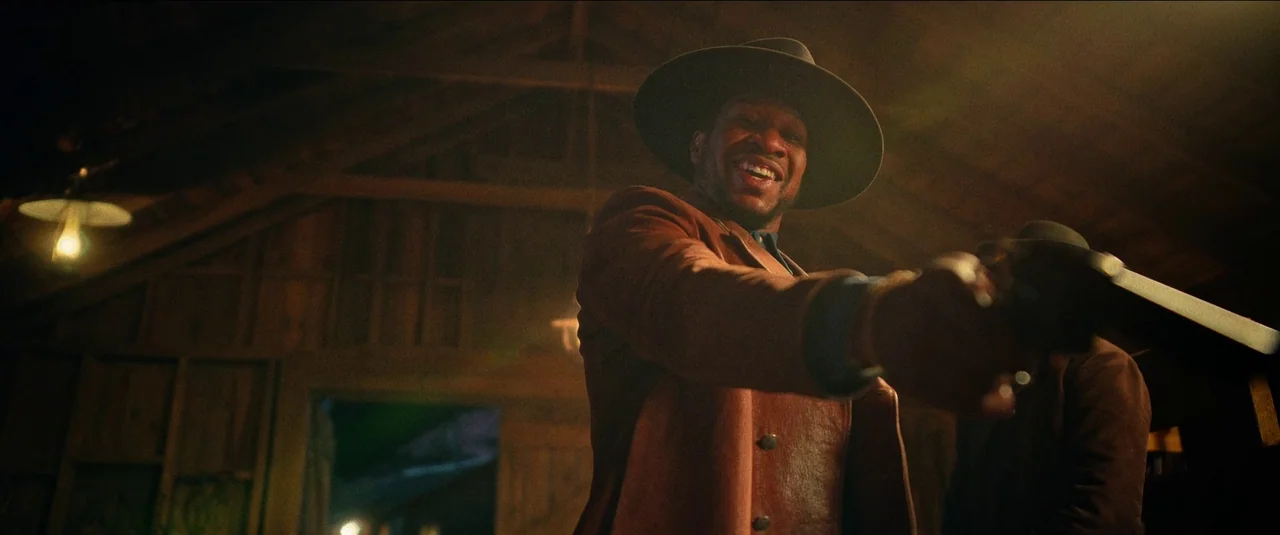 The Harder They Fall (2021) movie still: medium — A dimly lit wooden structure with rafters overhead and visible dust in the air. A man in a leather…