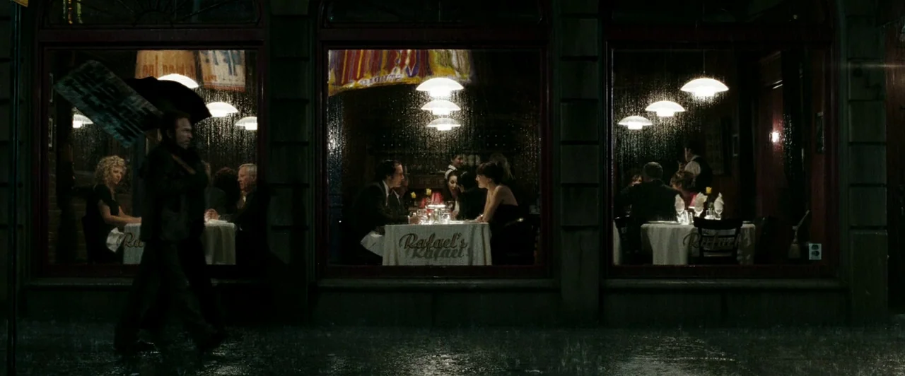 Watchmen (2009) movie still: wide — A street view of a restaurant exterior at night during a rainstorm. Patrons dine at white-clothed tables behind…