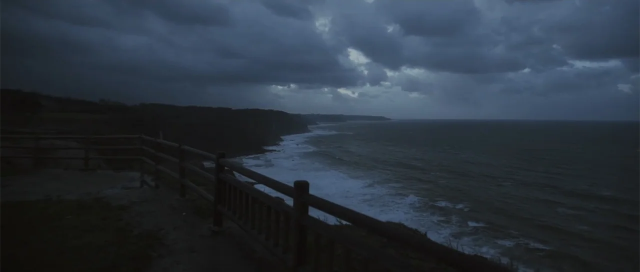 Taking Back the Waves (2017) movie still: establishing — A cliffside view overlooks a dark sea under a heavy cloud cover at night. A wooden fence…