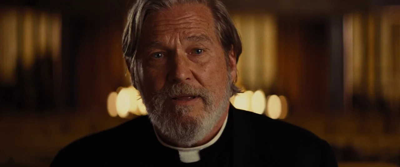 Bad Times at the El Royale (2018) movie still: closeup — A dimly lit room with a blurred background of distant light sources. A man in clerical…
