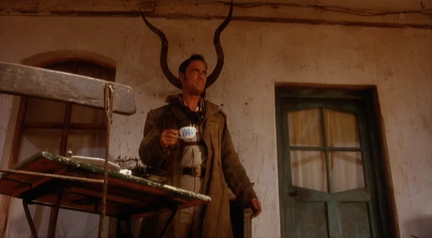 Dust Devil (1992) movie still: medium — A man stands on a weathered porch with light-colored stucco walls and a door. Large curved horns mounted on…