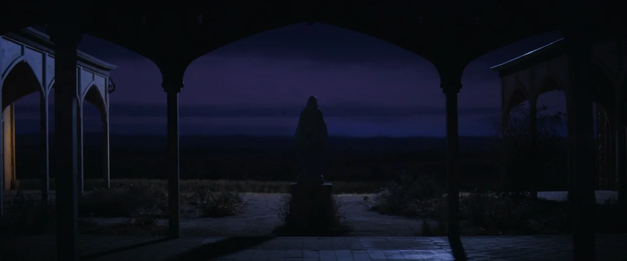 The New Boy (2023) movie still: wide — An open courtyard at dusk is framed by dark, arched wooden structural pillars. A stone statue of a religious…