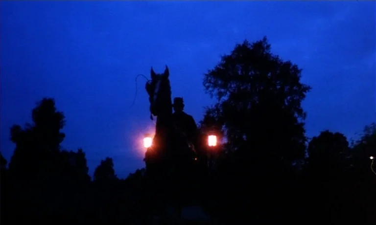 Katie Tippel (1975) movie still: wide — A dark carriage with two glowing lanterns travels along a road against a sky. Trees frame the subject in a…