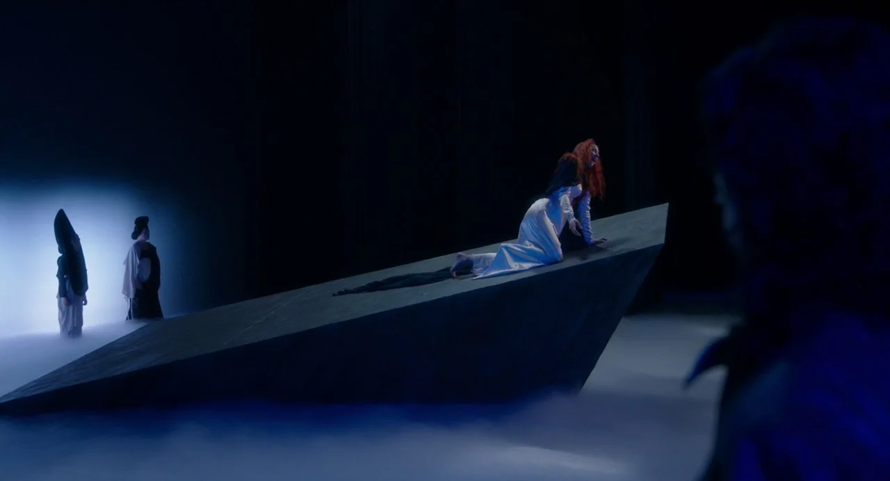 Annette (2021) movie still: wide — A dark theatrical stage features a large stone wedge surrounded by low-lying fog. A woman in a white dress moves…