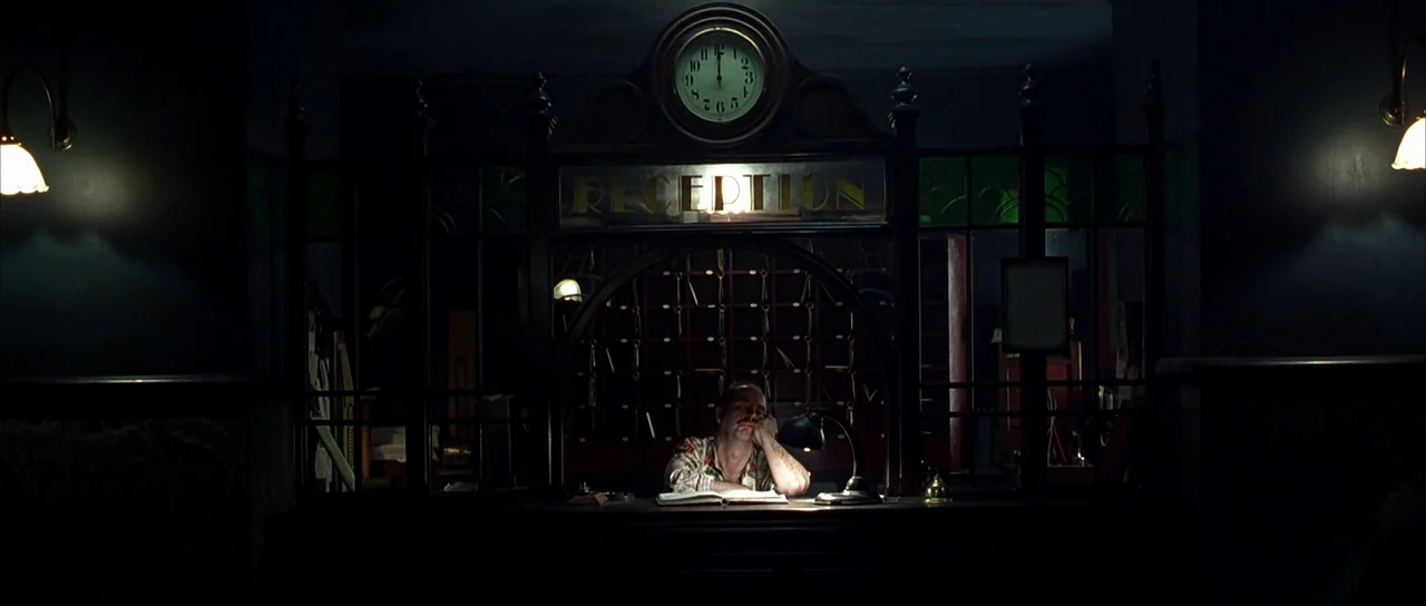 Dark City (1998) movie still: medium — A dimly lit hotel reception area features a wooden counter and a large clock. A lone clerk sits at the desk…