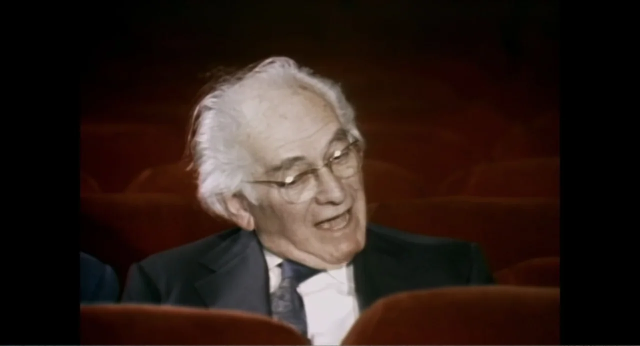 Made in England: The Films of Powell and Pressburger (2024) movie still: medium — An elderly man sits in a dimly lit, dark red movie theater seat.…